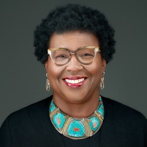 The image shows a smiling Black woman with short, curly hair and glasses. She is wearing a black top and a turquoise and coral necklace. Her earrings are gold. The background is a solid, muted gray. She appears to be in a studio setting.